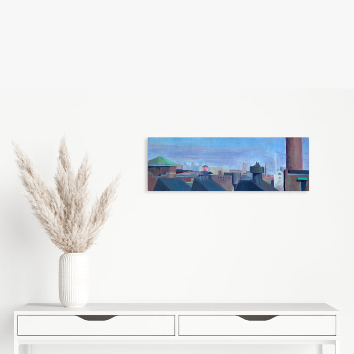 The artwork 'Long Rooftops' by Peter Colquhoun captures an urban skyline with traditional pitched rooftops against a soft blue sky. The oil painting features muted shades of gray and blue, depicting a series of connected buildings with chimneys and architectural details stretching across the horizontal canvas board, creating a serene cityscape perspective.