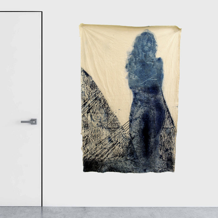 The artwork 'Crepuscular' by Johanna Ryan features a striking silhouette rendered in deep indigo cyanotype on coffee-stained canvas. Abstract textural patterns create a moody landscape effect beneath the ethereal figure. The 70x49 inch piece combines traditional photographic processes with mixed media elements, resulting in a dreamlike composition of blue tones against warm sepia background.