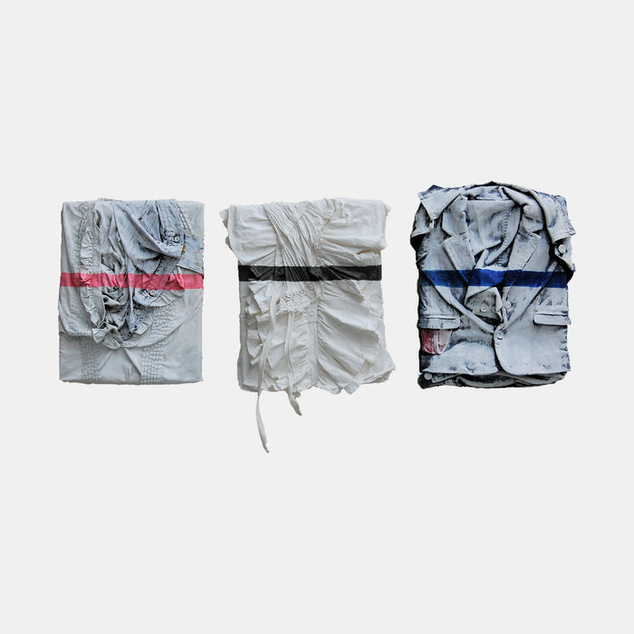 The artwork 'Family' by Noriko Okada features three folded fabric pieces arranged horizontally, each bound with different colored bands - red, black, and blue. The textured, wrinkled textiles in pale gray tones create a minimalist composition with subtle variations in light and shadow, painted in oil on fabric, measuring 13.5x11 inches each.