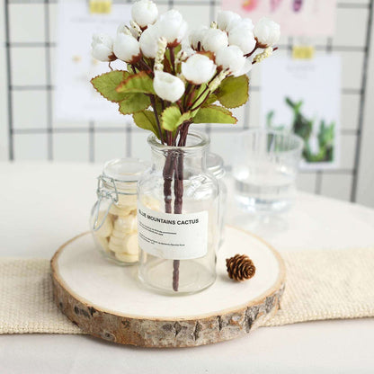 Artificial white flower arrangement in glass jar on wooden slice.