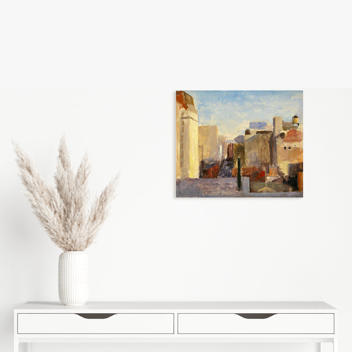 The artwork 'Seventeenth Street' by Peter Colquhoun depicts an urban cityscape with tall buildings in warm ochre and sandy tones against a soft blue sky. The oil painting on linen captures the architectural geometry of city structures, with subtle shadows and light playing across building facades, creating depth and atmosphere in an impressionistic style.
