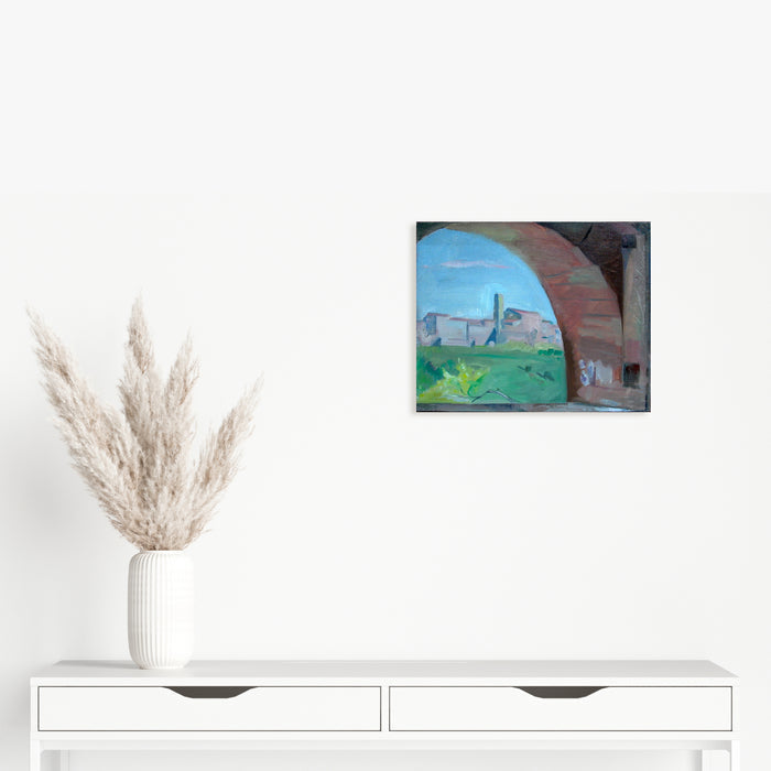 The artwork 'Through the Arch, Casole' by Peter Colquhoun captures a scenic Italian landscape viewed through a terracotta stone archway. The composition features vibrant blue skies, distant medieval buildings on a hilltop, and lush green countryside, all framed by the rustic arch's curved stonework. Oil on linen, 13x16 inches.