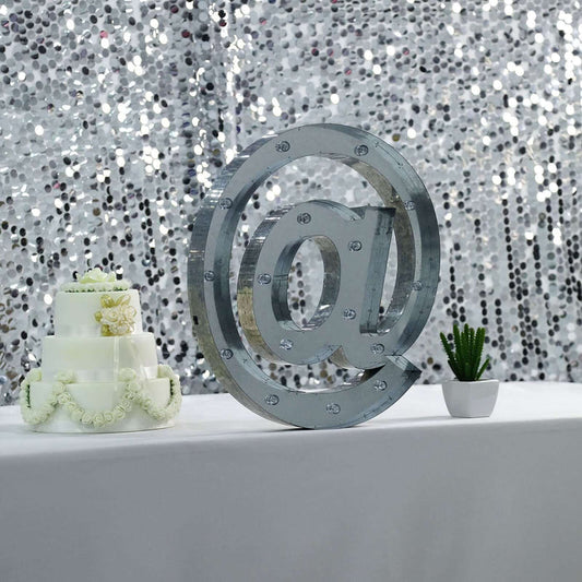 Decorative '@' sign with lights, cake, and plant on table against sparkling backdrop.